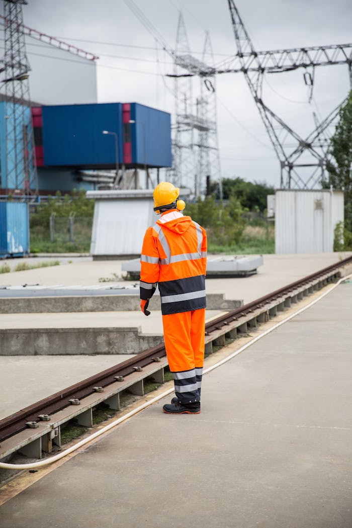 A construction worker in orange high-visibility gear stands at an industrial site with power lines.